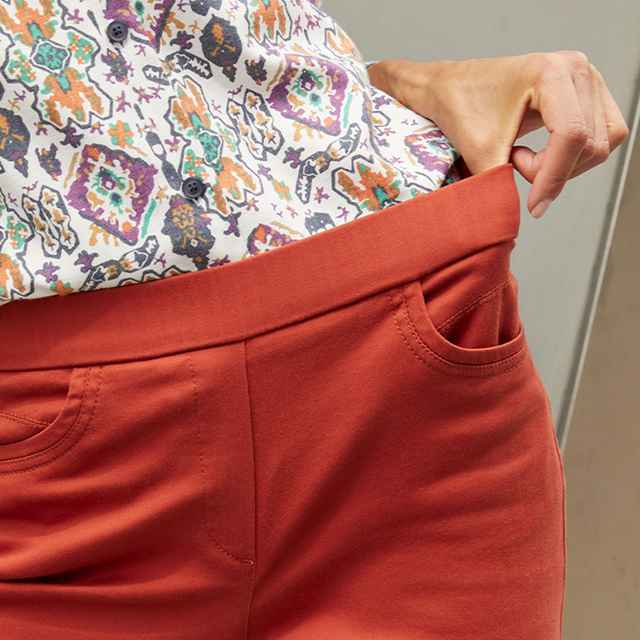Rote Hose mit elastischem Bund und Taschen, kombiniert mit einem bunt gemusterten Oberteil. Một Hand zieht am Hosenbund.