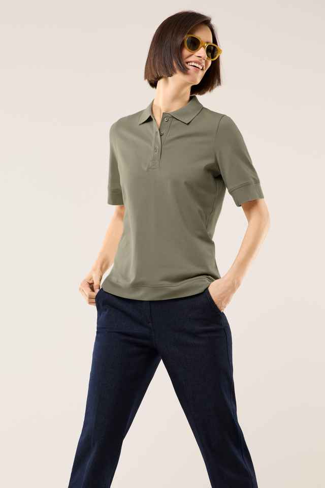 Frau mit kurzen Haaren trägt ein khaki Poloshirt, dunkle Hose und gelbe Sonnenbrille, lächelnd mit Händen in den Hosentaschen.