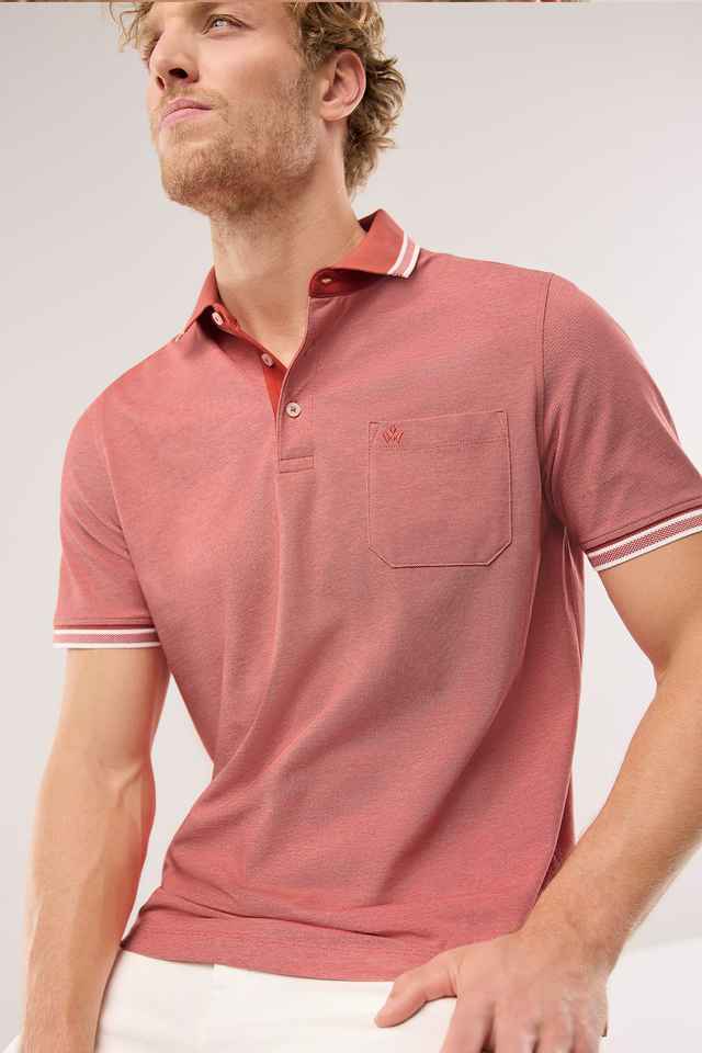 Mann in einem rosa Poloshirt mit Brusttasche und weißen Akzenten, sitzt entspannt und schaut zur Seite vor einem grauen Hintergrund.