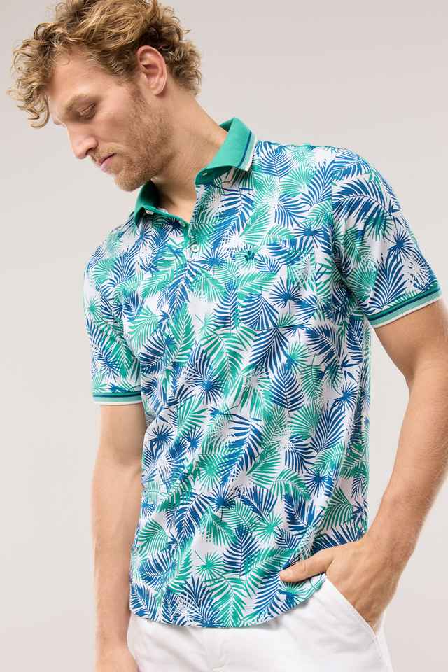 Mann mit lockigem Haar trägt ein buntes Polohemd mit tropischem Blätterdesign und eine weiße Hose.