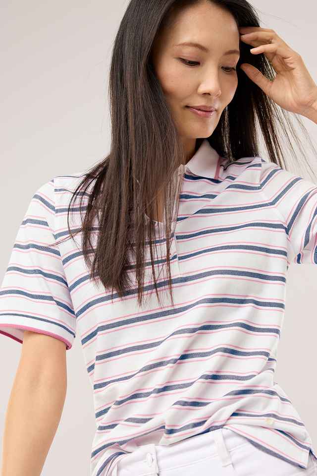 Frau mit langem, dunklem Haar trägt ein gestreiftes Poloshirt und weiße Hose, schaut nach unten und berührt ihr Haar.