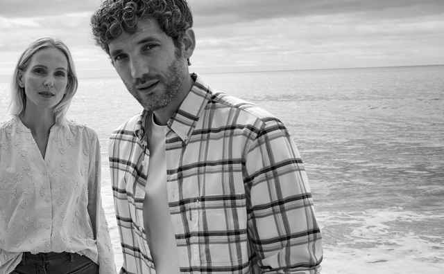 Ein Frau und ein Mann stehen am Strand. Sie trägt eine bestickte Bluse und er ein offenes kariertes Hemd mit einem weißen Shirt darunter.