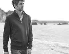 Ein Mann geht am Strand entlang und trägt einen Troyer, darunter ein gestreiftes Hemd und dazu eine Jeans.