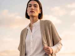 Frau in weißer Bluse und beige Strickjacke steht im Freien bei Sonnenuntergang, schaut selbstbewusst in die Kamera.