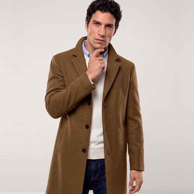 Brauner Mantel über einem cremefarbenen Pullover und blau-weiß gestreiftem Hemd, kombiniert mit dunkler Jeans. Der Stil ist klassisch und elegant.