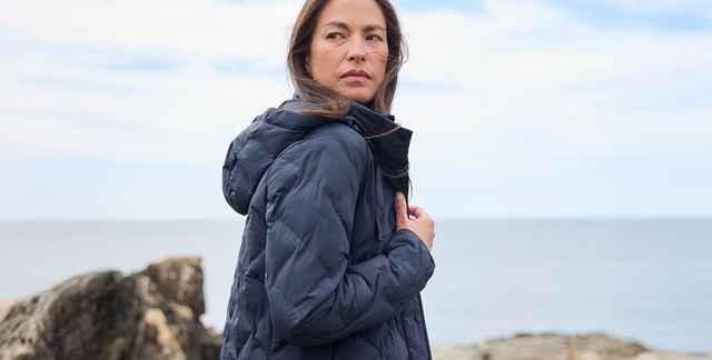Frau mit dunkelblauer Jacke steht am Meer, schaut nach rechts. Hintergrund: Felsen und Himmel.