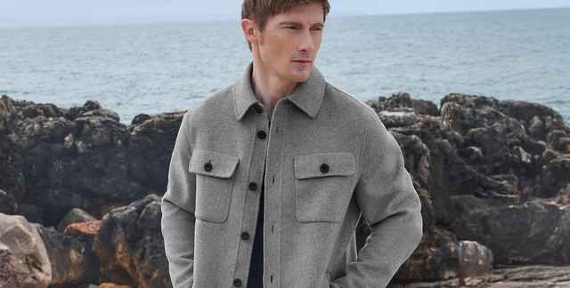 Mann in grauer Jacke steht vor felsiger Küste mit Blick aufs Meer.