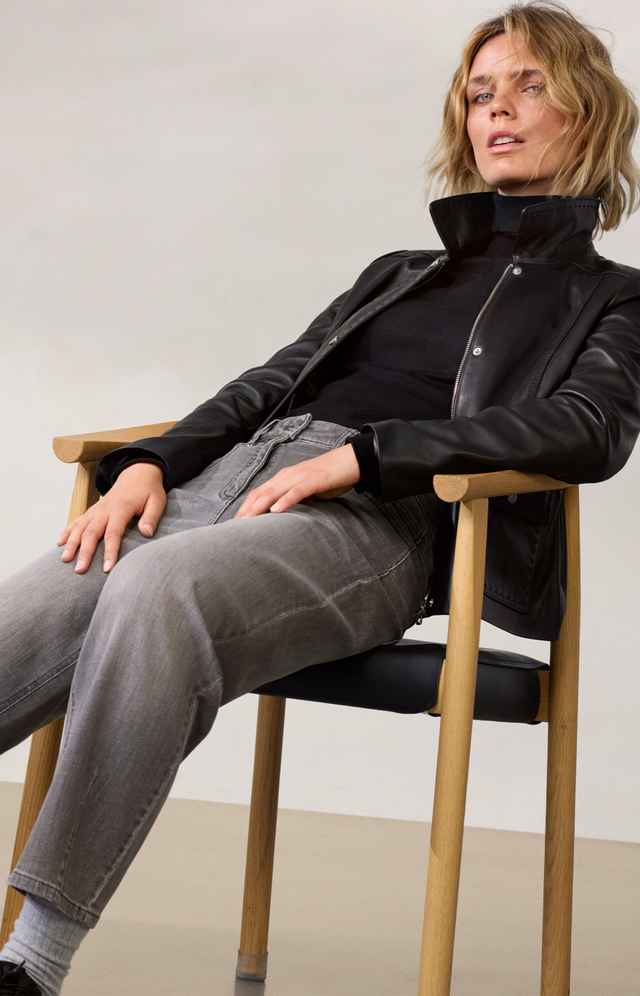 Frau im schwarzen Rollkragenpullover und Lederjacke sitzt entspannt auf einem Stuhl. Sie trägt graue Jeans und hat blondes Haar.