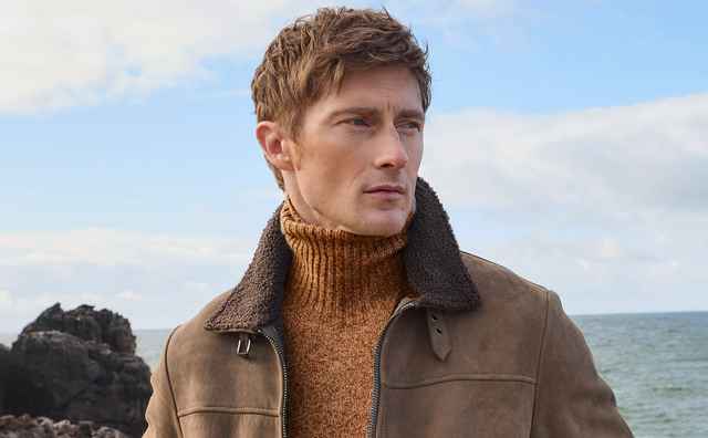 Mann in brauner Jacke und Rollkragenpullover steht auf felsiger Küste, blickt seitlich aufs Meer.