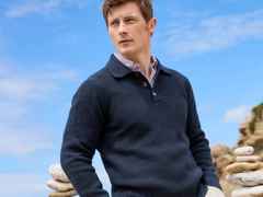 Ein Mann in einem dunkelblauen Pullover steht im Freien vor einem Stapel Steine, mit blauem Himmel im Hintergrund.