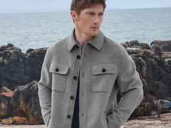 Ein Mann in grauer Jacke steht vor einer felsigen Küstenlandschaft mit Meer im Hintergrund.