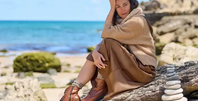 Frau in beigem Pullover sitzt nachdenklich auf einem Felsen am Strand, Stapel kleiner Steine neben ihr, Meer im Hintergrund.