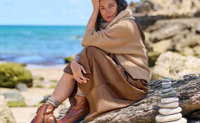 Frau in beiger Kleidung sitzt nachdenklich auf einem Felsen am Strand, im Hintergrund sieht man das Meer und blauen Himmel.