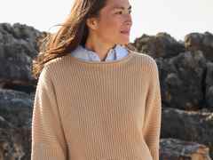 Frau in beigefarbenem Strickpullover steht draußen vor Felsen, schaut zur Seite.