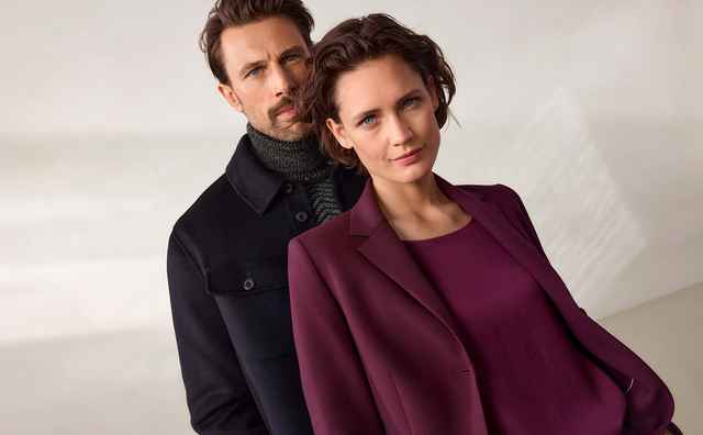 Ein Mann und eine Frau in eleganter Kleidung stehen nebeneinander, vor einem neutralen, hellen Hintergrund.