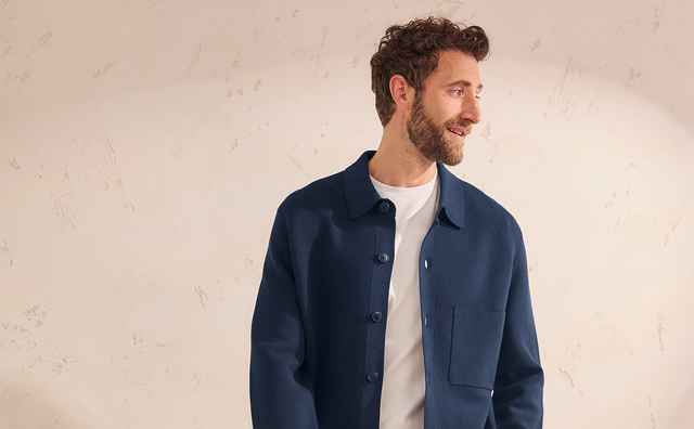 Ein Mann trägt eine blaue Jacke und graue Hose, steht lächelnd vor einem beigen Hintergrund.