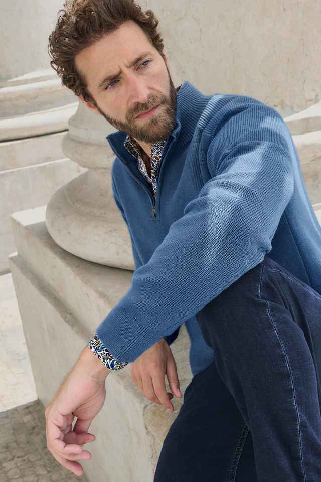 Mann mit Bart sitzt nachdenklich in blauer Jacke und Jeans auf einer Steintreppe.