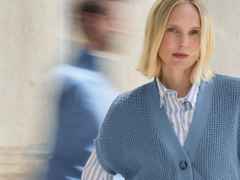 Blonde Frau in blauem Pullover und gestreifter Bluse, lächelnd. Verschwommene, sich bewegende Person im Hintergrund.