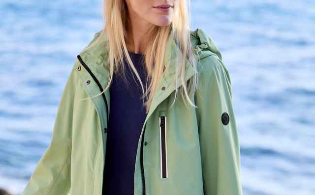 Frau in hellgrüner, sportlich-eleganter Regenjacke mit Kapuze und schwarzen Reißverschlüssen über einem dunkelblauen Oberteil.