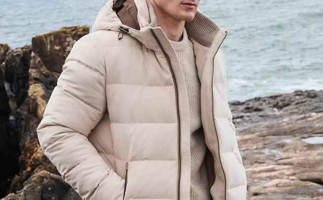 Mann in einer hellbeigen, wattierten Outdoorjacke mit Kapuze über einem grob gestrickten Pullover – sportlich-warmer Stil.