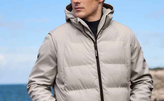 Mann in einer hellgrauen Outdoorjacke mit Kapuze, schwarzem Reißverschluss und sportlich-funktionalem Stil.