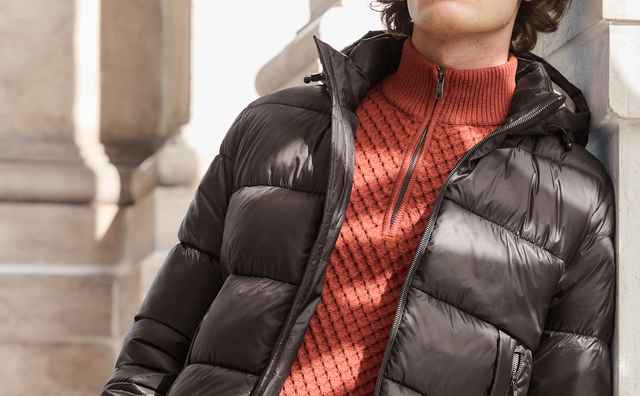 Mann in einer schwarzen, glänzenden Steppjacke mit Kapuze und orangefarbenem Strickpullover – sportlicher Winterstil.