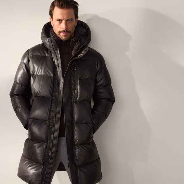 Mann in einer schwarzen, glänzenden Steppjacke mit Kapuze über einem dunklen Strickpullover – markanter, sportlicher Winterstil.