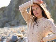Frau mit braunen Haaren trägt gestrickten Pullover, steht auf einem steinigen Strand und hält ihre Hand schützend vor die Sonne.