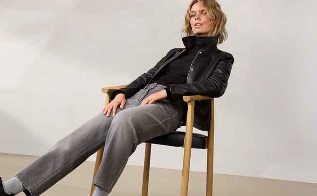 Eine Frau in schwarzer Lederjacke und grauen Jeans sitzt lässig auf einem Stuhl vor einem neutralen Hintergrund.