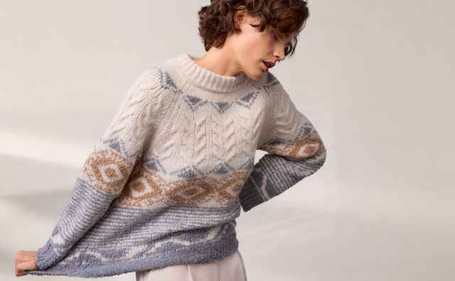 Person mit lockigem Haar trägt einen gestrickten Pullover mit geometrischem Muster in Creme, Blau und Braun vor einem neutralen Hintergrund.