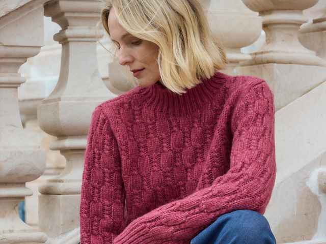 Person mit blonden Haaren und rotem Strickpullover sitzt vor einer dekorativen Steintreppe.