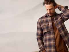 Ein Mann trägt ein karriertes Hemd über einem braunen Pullover und blickt leicht nach unten, eine Hand hinter dem Kopf.