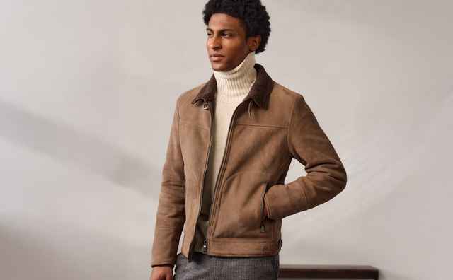 Ein Mann in einer braunen Jacke und Rollkragenpullover steht mit einer Hand in der Tasche, vor einem minimalistischen Hintergrund.