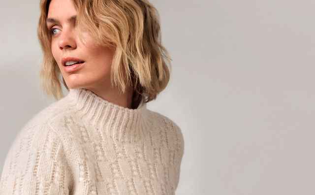 Frau mit blonden Haaren trägt einen cremefarbenen Strickpullover und Hose, sitzt auf einem Stuhl und blickt zur Seite.